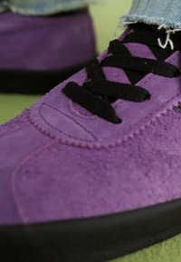 Sapatilhas de camurça roxa com atacadores pretos, apresentando uma textura suave e uma sola de borracha preta em contraste. Close-up destacando os detalhes das costuras.