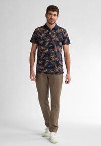 Polo navy con motivo floreale in beige, abbinato a pantaloni marrone chiaro e sneakers bianche. La camicia ha un colletto e chiusura con bottoni.