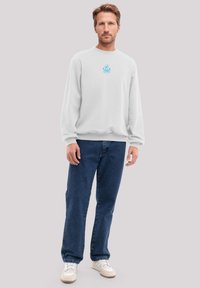 White sweatshirt with a blue graphic design, round neckline, and ribbed cuffs, paired with dark denim jeans and white sneakers.