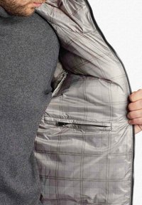 Uomo che indossa un maglione grigio apre una giacca a scacchi grigia mostrando una tasca con zip interna sul lato destro.