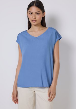 Haut bleu à manches courtes avec un col en V, présentant des accents de dentelle texturée sur les épaules et une coupe décontractée. Ourlet avant légèrement arrondi.