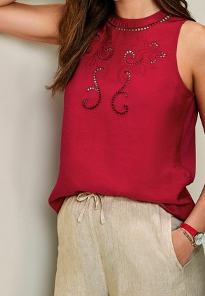 Kvinde iført en rød ærmeløs top med blomsterbroderede udskæringer og beige bukser med snor, med et rødt ur og armbånd på håndleddet.