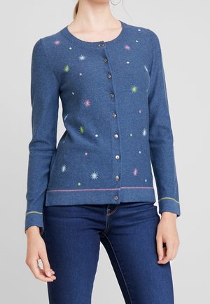 Blauw gebreid vest met knopen en kleurrijke kleine zonnestraal- en stippenpatronen, gecombineerd met donkerblauwe jeans bij een staande vrouw.