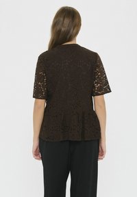 Naine on nähtud seljaga, kandes lühikeste varrukatega tumeda pruunika lillemustriga pitsist toppi, millel on peplum-äär ja mustad püksid.