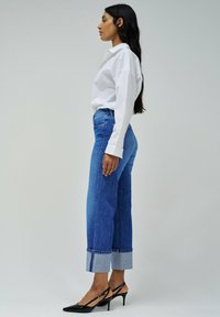 Camisa blanca de cuello con jeans de mezclilla azul de cintura alta, con puños enrollados y tacones negros de punta. Composición en vista lateral.