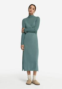 Lång, figurnära klänning i mjukt teal-tyg med ribbat mönster, hög krage och sidoklipp. Bärs med beige slip-on-skor och krämfärgade strumpor.