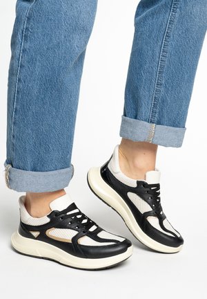 Person wearing black and white mesh sneakers with thick soles and cuffed blue jeans on a white background.