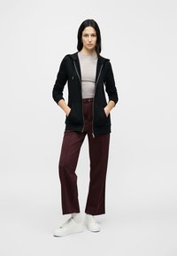 Zwarte zip-up hoodie met een zachte afwerking boven een lichtgrijze nauwsluitende top; gecombineerd met bordeauxrode wijde broekspijpen en witte sneakers.