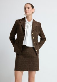 Brązowa kurtka z tweedu z złotymi guzikami, zestawiona z białą bluzką z detalem w postaci wiązania oraz pasującą brązową mini spódniczką z tweedu.