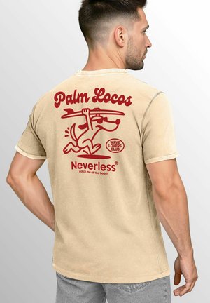 Beige T-Shirt aus Baumwolle mit leicht zerknitterter Textur. Es hat ein rotes Grafikdesign eines Hundes, der ein Surfbrett hält, und Text.