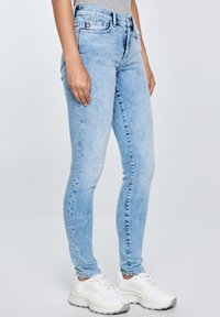 Inimene, kes kannab helesiniseid kitsaid teksapükse ja valgeid tossusid, seisab ühevärvilise helehalli tausta ees.