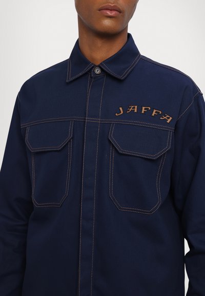 Camicia in denim blu navy con cuciture marroni, due tasche sul petto e 'JAFFA' ricamato in corsivo sul lato sinistro. Chiusura con colletto a bottoni.