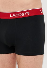 Boxer shorts noirs avec une taille rouge comportant le texte "LACOSTE" en blanc. Tissu lisse avec un léger brillant, design ajusté et sans motifs visibles.