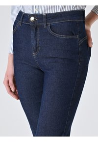 Primo piano di una persona che indossa jeans in denim blu scuro con cuciture visibili, bottone in metallo e tasche anteriori, mano appoggiata sui fianchi.