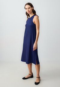 Navy mouwloos jurkje met een geribbelde textuur, knielange ontwerp en een zijsplit. Gecombineerd met zwarte platte schoenen met enkelbandjes.