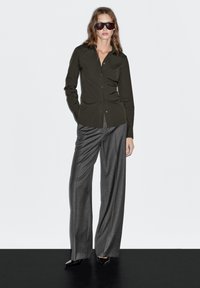 Olive button-up shirt paired with gray wide-leg trousers. Model wears dark sunglasses and black pointed-toe heels. Smooth fabric texture.