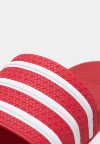 Κοντινό πλάνο κόκκινου λουριού σανδαλιού με υφή στην επιφάνεια και τρεις παράλληλες λευκές ρίγες πάνω του.