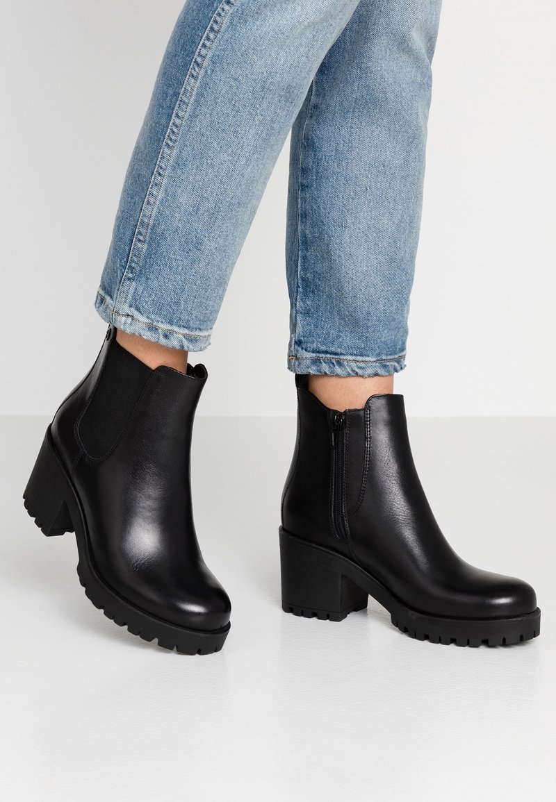 Personne portant un jean bleu clair et des bottines en cuir noir à talons épais et fermetures éclair latérales sur un sol blanc.