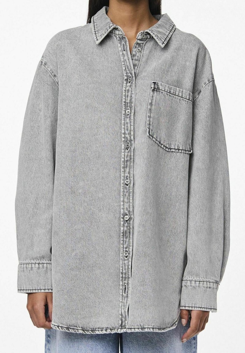 Femme portant une chemise en jean gris clair oversize boutonnée avec une seule poche poitrine, associée à un jean bleu.