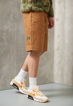Mann trägt braune Shorts mit blauem "H"-Logo, weiße Sportsocken und beige Sneakers mit orangefarbenem Nike-Swoosh, steht auf grauem Boden.
