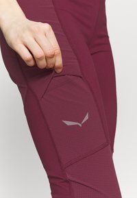 Mörkröda träningsleggings med texturerat tyg och en åtsittande design, med en diskret grå logotyp nära knäet och en sidoficka.