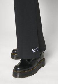 Svarta bomullsshorts med vida ben och "AIR"-logotyp i vitt, matchat med blanka svarta platåskor med gula sömmar.