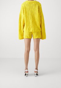 Gul stickad tröja med texturerat mönster, kombinerad med matchande shorts. Modellen bär vita klackar. Bakgrunden är enkel.