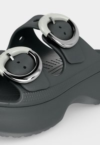 Σκούρα γκρι σανδάλι τύπου slip-on με δύο ρυθμιζόμενους φαρδιούς ιμάντες, ο καθένας με μια μεγάλη ασημένια και λευκή κυκλική αγκράφα.