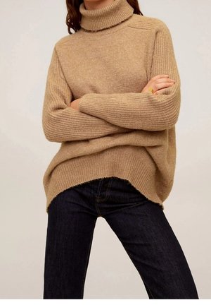 Kvinde iført en beige højhalset striktrøje med ribstrikkede ærmer og mørkeblå jeans, armene krydset over brystet.