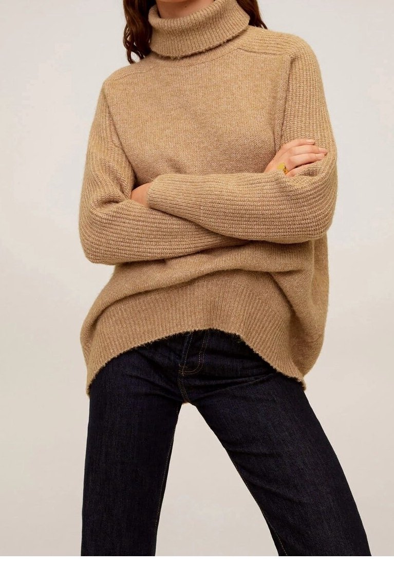 Femme portant un pull en maille beige à col roulé avec des manches côtelées et un jean bleu foncé, les bras croisés sur la poitrine.