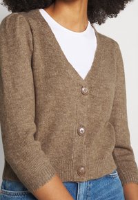 Hnědý pletený kardigan s hlubokým výstřihem do V a třemi knoflíky, s žebrovanými manžetami a lemem. Měkká struktura a přiléhavý střih.