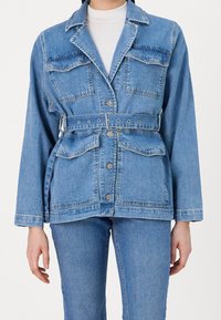 Giacca in denim di colore azzurro chiaro, con due tasche sul petto, una vita con cintura e colletto classico. Bottoni lungo la parte anteriore. Maniche lunghe.