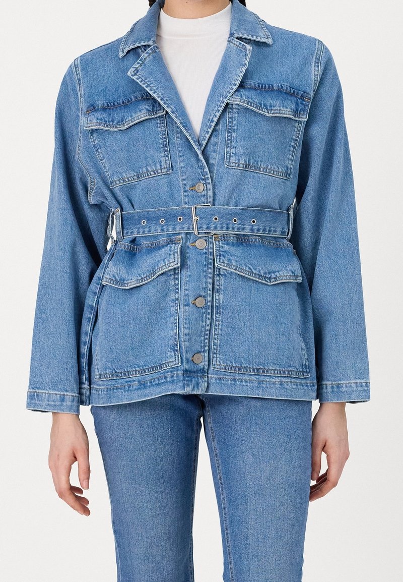 Giacca in denim di colore azzurro chiaro, con due tasche sul petto, una vita con cintura e colletto classico. Bottoni lungo la parte anteriore. Maniche lunghe.