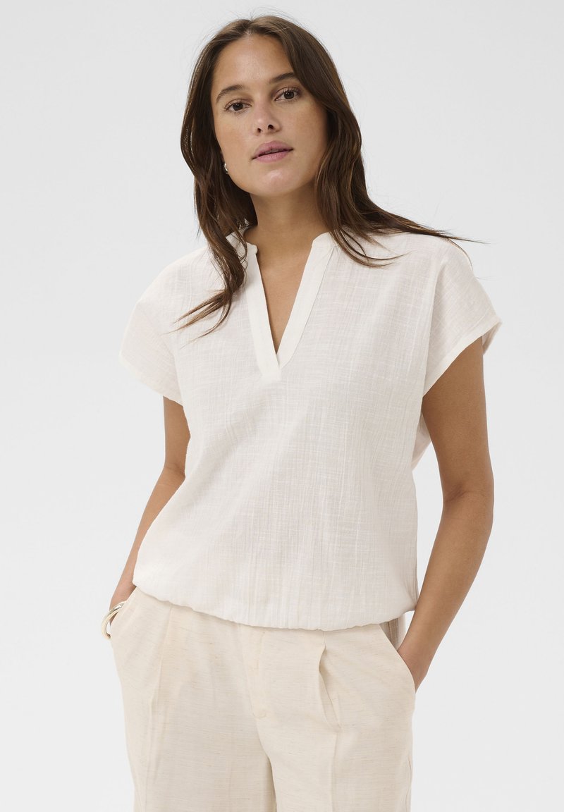 Femme aux longs cheveux bruns portant un chemisier blanc à manches courtes et un pantalon beige clair, les mains dans les poches, fond neutre.