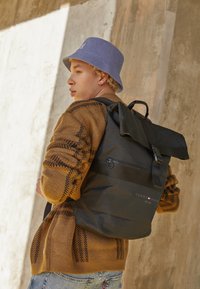 Sac à dos noir à rabat avec une poche zippée, fabriqué en matériau texturé. Porté avec un pull brun à motifs et un chapeau seau bleu clair.