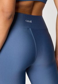 Μπλε κολάν με ψηλή μέση, από λείο ύφασμα, προσφορά ελαστικής ζώνης και διακριτική επωνυμία. Σχεδιασμένα για άνεση και  ευελιξία στην αθλητική ένδυση.