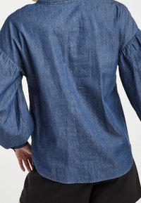 Denimskjorta med samlade ballongärmar, mörkblå färg, mjuk textur och rundad fåll. Bakifrån, inga synliga knäppningar.