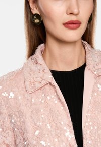 Helle rosa Spitzenjacke mit Pailletten, ausgestattet mit einem kragenartigen Hals und zartem Blumenmuster. getragen über einem schwarzen strukturierten Oberteil. Runde goldene Ohrringe.