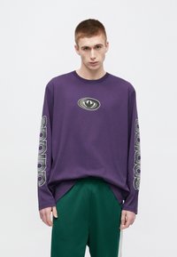 Violeta T-krekls ar garām piedurknēm un ovālu logotipu, uz piedurknēm baltā krāsā attēlots "sp". Valkāts kopā ar zaļām biksēm, uzsverot ērtu piegriezumu un dizainu.