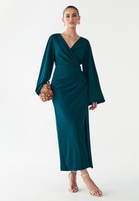 Vestido cruzado color azul verdoso con mangas largas, detalle fruncido en el costado y silueta fluida. Combinado con un clutch dorado con lentejuelas y sandalias beige de tiras.