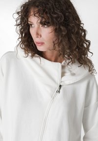 Sudadera blanca con cremallera hecha de un tejido suave, que presenta una gran capucha y una cremallera lisa. Diseño simple sin patrones o acentos visibles.