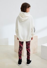 White oversized hoodie with a hood and elastic hem, paired with black leggings featuring pink abstract lines, and black high-top sneakers.