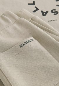 Bež bombažna majica z izrazitim črnim logotipom in vezenim napisom "ALLSAINTS" na strani. Gladek otip in sproščen kroj.
