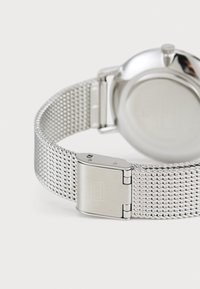 Reloj de pulsera de malla plateada con una caja de metal pulido. Cuenta con una correa de malla y un cierre tipo de sobre, mostrando una superficie lisa y reflectante.