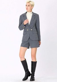 Blazer à carreaux bleu marine et blanc avec double boutons, associé à un short assorti. Des bottes en cuir noires jusqu'aux genoux complètent la tenue.