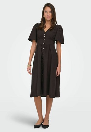 Žena stojaca v čiernych midi šatách s naberanými rukávmi, zapínanie na gombíky vpredu, doplnené čiernymi špicatými balerínkami a zlatým náramkom.