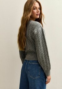 Maglione grigio a maglia con vita aderente e maniche testurizzate, abbinato a jeans blu a vita alta, visto da dietro.