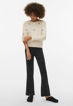 Maglione beige lavorato a maglia con piccoli motivi a cuore, abbinato a pantaloni neri a zampa con linee ondulate argentate. Scarpe nere slip-on.