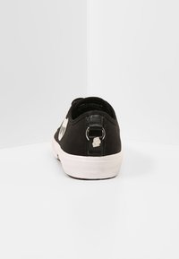 Musta matalavartinen tenniskenkä valkoisella kumipohjalla, takana vetonauha metallirenkaalla ja pieni hopeinen logo, asetettuna valkoista kulmaa vasten.