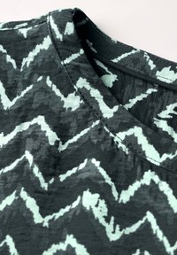 Schwarzer Stoff mit einem Zickzackmuster in hellem Minzgrün. V-Ausschnitt-Design mit gesticktem Ausschnittdetail. Weiches, texturiertes Material.